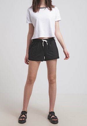 Femme portant un t-shirt court blanc, un short noir à cordon et des sandales plateformes noires, debout devant un fond clair uni.