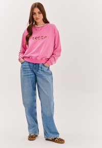 Różowa bluzka z haftem przedstawiającym postać z kreskówki, w połączeniu z jasnoniebieskimi dżinsami o szerokich nogawkach i butami slip-on w zwierzęcy wzór.
