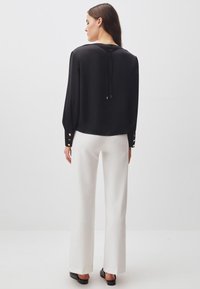 Camicia nera a maniche lunghe con scollatura a V e dettaglio del nodo sul retro, abbinata a pantaloni bianchi a gamba larga e scarpe nere. Tessuto morbido e leggero.