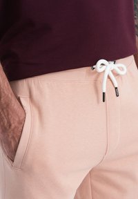Světle růžové tepláky s jemnou texturou, se bočními kapsami a bílou šňůrkou s černými koncovkami. Nosené s vínovým tričkem.