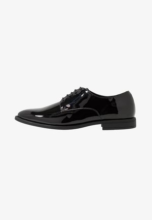 Chaussure de ville pour homme en cuir verni noir avec fermeture à lacets et talon bas empilé, présentée en profil latéral sur fond blanc.
