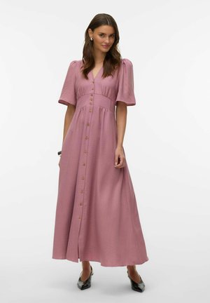 Femme portant une robe midi mauve boutonnée à l'avant avec des manches courtes bouffantes et des escarpins noirs à bouts pointus, debout sur un fond blanc.