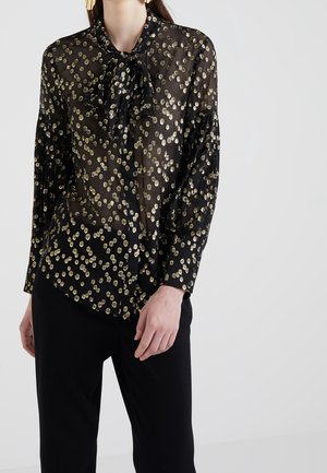 Kobieta ubrana w przezroczystą czarną bluzkę w złote groszki z kokardą, w zestawie z czarnymi spodniami na jednolitym tle.