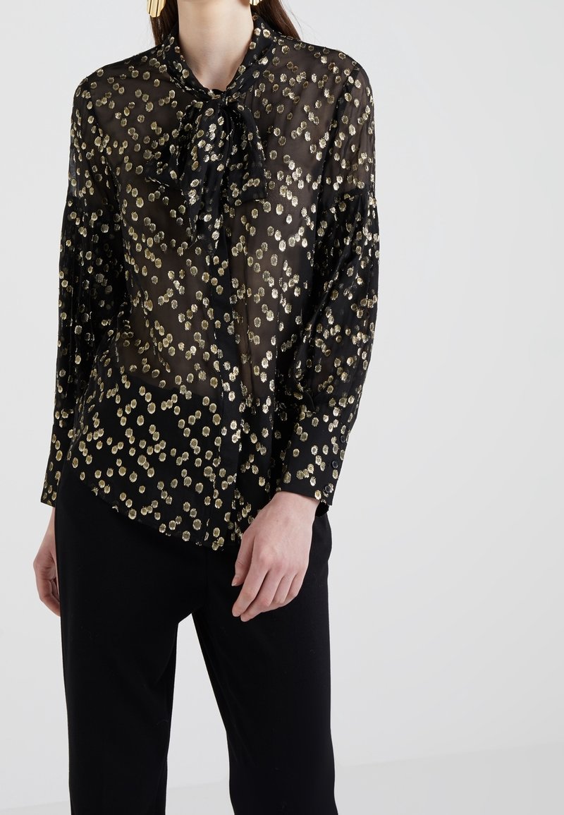 Femme portant un chemisier noir transparent à pois dorés avec un nœud papillon, associé à un pantalon noir sur un fond uni.