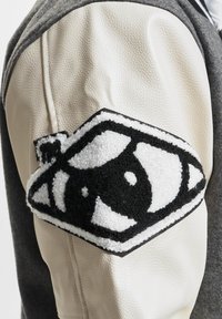 Grå og hvit jakke med lærermuffer og et teksturert svart-hvitt-patch med et stilisert øye-design på overarmen.