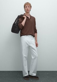 Bruine lange mouwen polo shirt, witte wijde broek en donkere schoenen. Model draagt een donkere tas, tegen een lichtgrijze achtergrond.