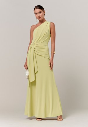 Kobieta w jasnozielonej wieczorowej sukni na jedno ramię z marszczonym pasem, trzymająca małą białą kopertówkę, nosząca złotą bransoletkę i kolczyki, stojąca.