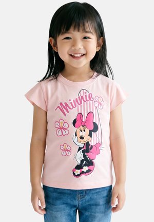 Fille souriante aux cheveux noirs portant un t-shirt rose Minnie Mouse et un jean bleu, debout devant un fond blanc uni.