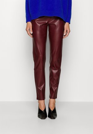 Osoba ubrana w bordowe skórzane spodnie, czarne szpilki z ostrym czubkiem oraz królewsko niebieski sweter, stojąca na tle jasnego, jednolitego tła.