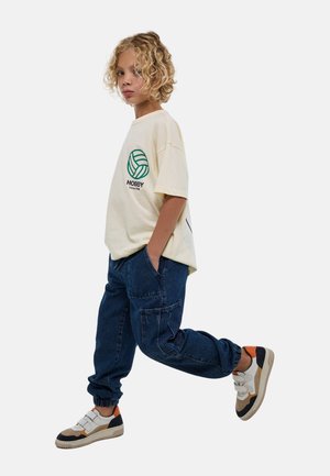 T-shirt en crème pour adolescents avec un graphique de ballon de volley-ball circulaire vert et le texte "HOBBY", associé à un pantalon cargo bleu foncé et des baskets multicolores.