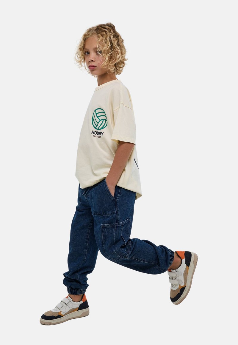 T-shirt en crème pour adolescents avec un graphique de ballon de volley-ball circulaire vert et le texte "HOBBY", associé à un pantalon cargo bleu foncé et des baskets multicolores.