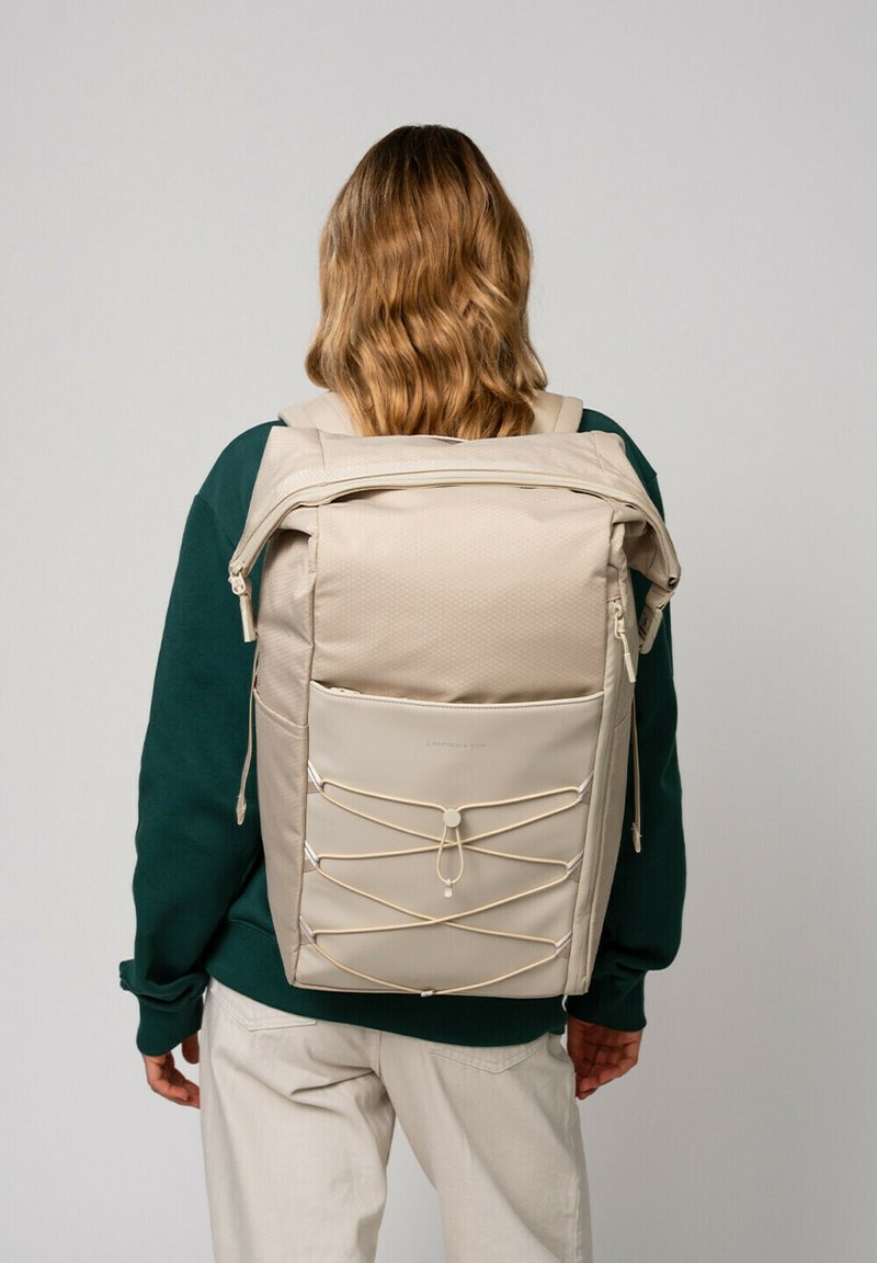 Sac à dos beige avec une surface texturée, dotée d'une poche avant et de détails en cordon élastique. Porté sur le dos, avec des bretelles réglables.