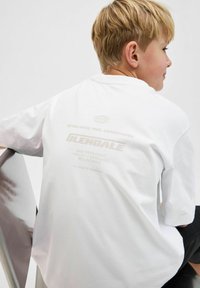Balta kokvilnas T-kreklu ar īsām piedurknēm. Aizmugurē redzami gaiši brūni printēti teksti un logotipi, tostarp "Iegādāts 1990, Kopenhāgena."