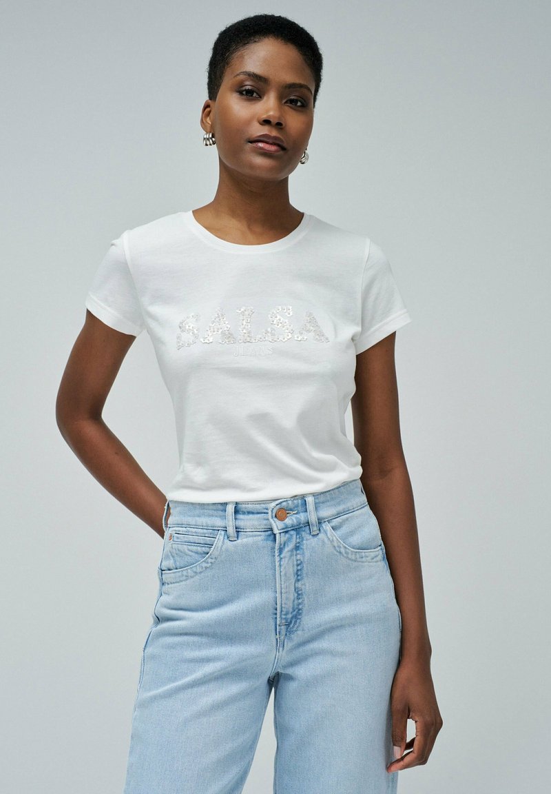 Camiseta blanca de algodón con "S.A.L.S.A." en brillantes lentejuelas, combinada con unos jeans de tiro alto en azul claro. Diseño de cuello redondo simple.