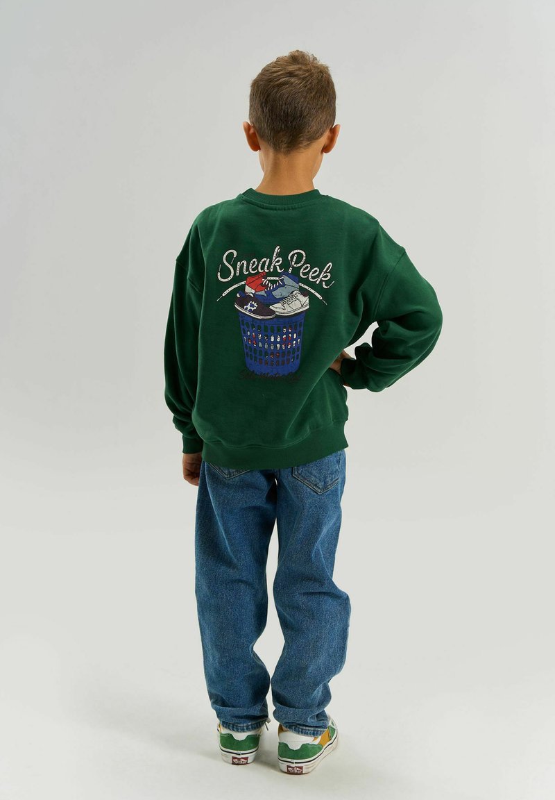 Garçon aux cheveux courts, de dos, portant un sweat-shirt vert avec l'inscription « Sneak Peek » et un motif de basket, un jean bleu et des baskets vertes.