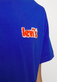 Mėlynas medvilnės marškinėliai su tekstūruotu oranžinės ir baltos spalvos "levi's" logotipu kairėje krūtinės pusėje. Trumpomis rankovėmis ir apvalia iškirpte.