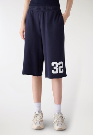 Persona che indossa pantaloncini blu navy alla lunghezza del ginocchio con il numero 32 stampato sulla gamba sinistra, calzini bianchi e sneakers bianche, in piedi con le mani nelle tasche.