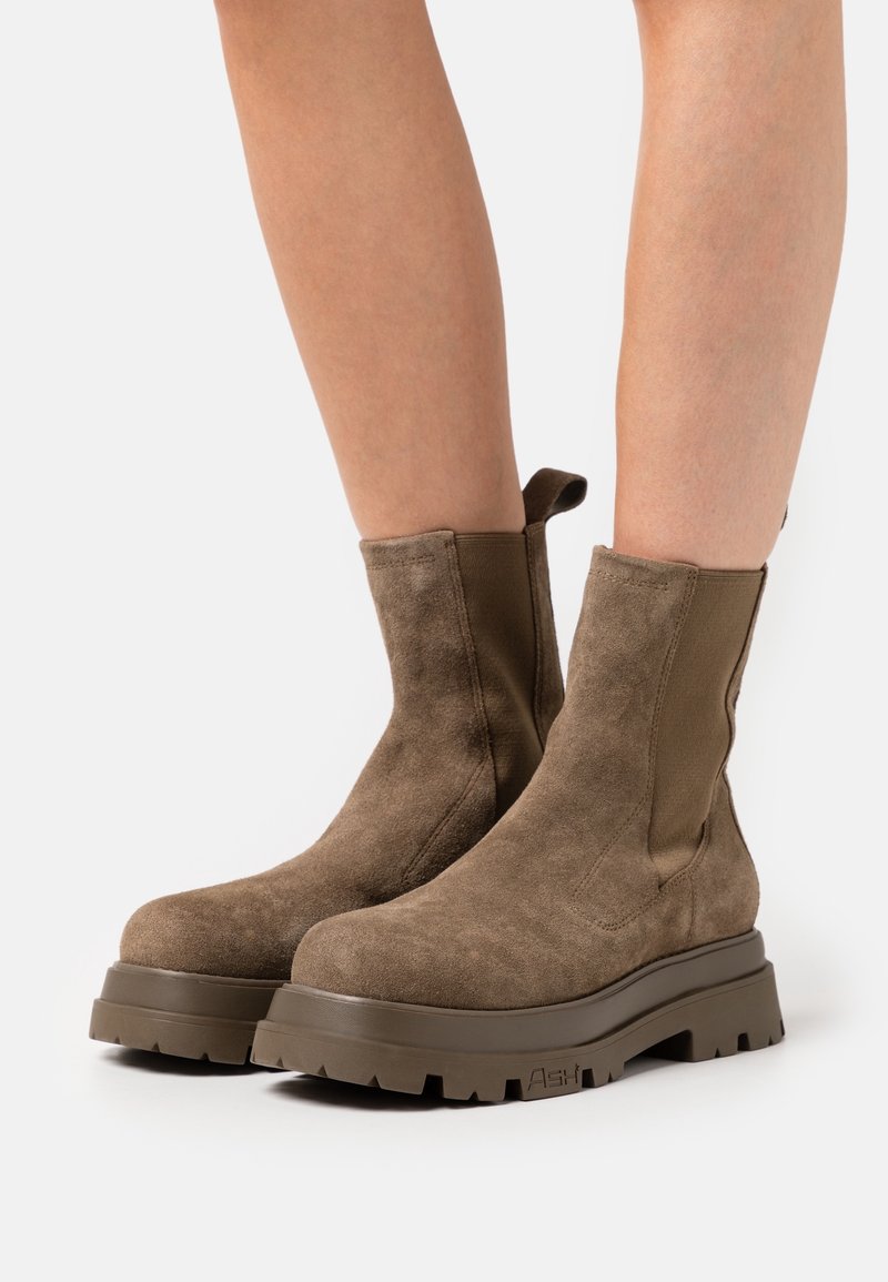 Bottines en daim marron avec semelles épaisses crantées portées sur des jambes nues sur un fond blanc uni.