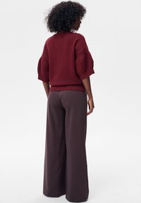 Bordowy sweter na drutach z krótkimi bufkami, noszony z szerokimi, ciemnobrązowymi spodniami o luźnym kroju, podkreślający fakturę tkanin i swobodny fason.