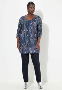 Marineblaue lange Tunika mit lila und pinkem abstraktem Muster, ausgestattet mit dreiviertel Ärmeln. Kombiniert mit schwarzen engen Hosen und silbernen Ankle-Boots.