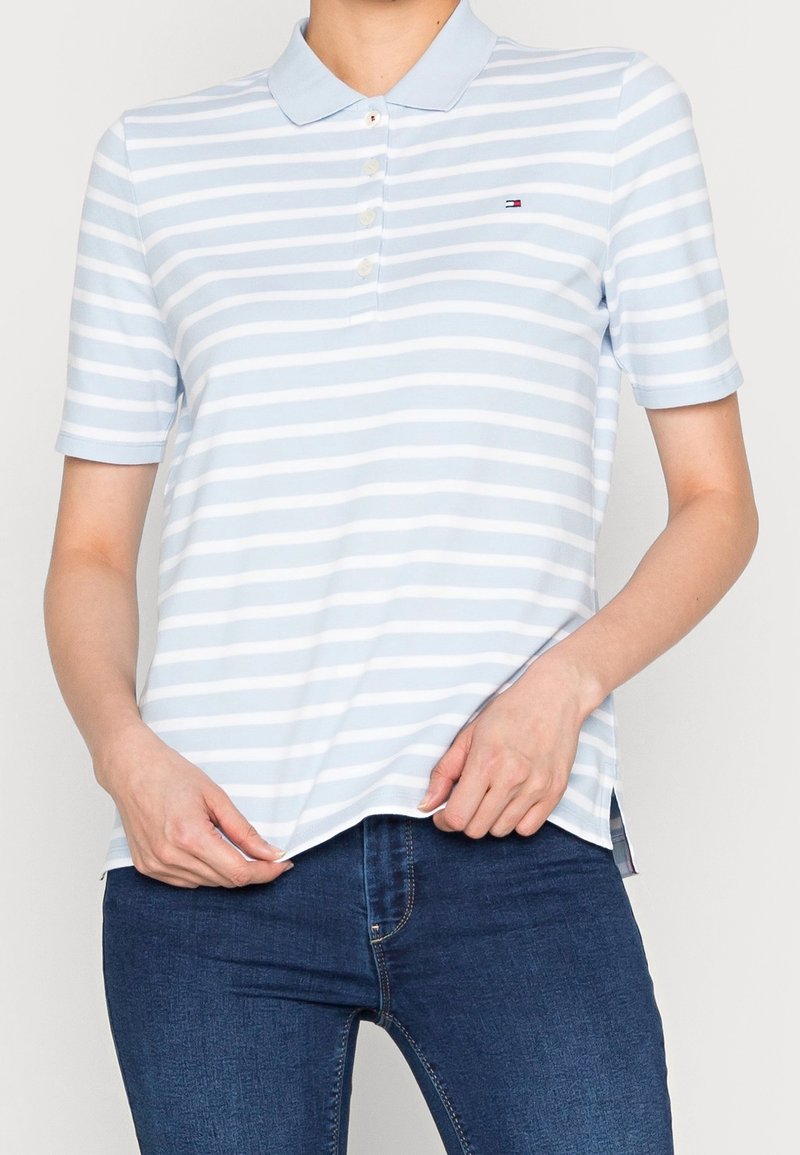 Femme portant un polo à manches courtes rayé bleu clair et blanc, et un jean bleu foncé, tenant l'ourlet de la chemise avec les deux mains.
