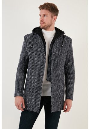 Cappotto strutturato di colore blu navy con cappuccio nero, dotato di chiusura a bottoni e due tasche frontali. Indossato sopra un maglione bianco.