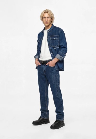 Jeune homme aux cheveux blonds portant une veste en denim bleu foncé et un jean avec des chaussures noires, debout les mains dans les poches sur fond blanc.