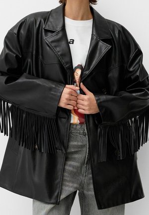 Personne portant une veste en cuir noire avec des détails à franges sur les manches, par-dessus un t-shirt blanc graphique et un jean gris, mains ajustant le devant de la veste.