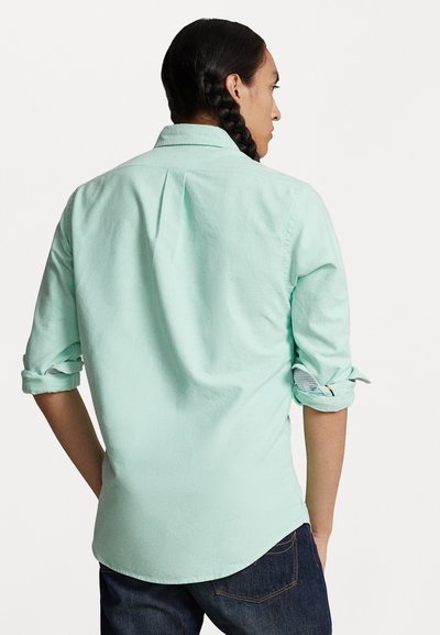 Light mint green button-up shirt with rolled sleeves, featuring a back pleat and a curved hem, paired with dark jeans.