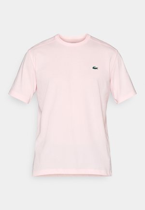 Gaiši rozā īsām piedurknēm kreklis ar apaļu kakla izgriezumu, uz kura kreisās krūts ir neliels zaļš krokodila logo uz vienkārša fona.