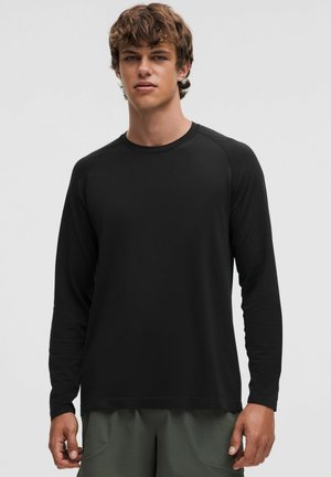 Jeune homme portant une chemise noire ajustée à manches longues et un pantalon vert olive, debout devant un fond clair uni.