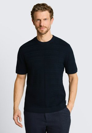 Uomo con capelli castano chiaro che indossa una camicia nera a maniche corte dalla trama evidente e pantaloni scuri a righe sottili, in piedi con una mano in tasca.