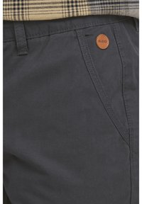 Pantaloni gri închis din țesătură împletită, cu un buzunar standard ce poartă un eticheta circulară maro "BLEND". Textură netedă cu cusături curate.
