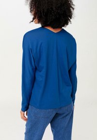 Person med lockigt hår som bär en blå långärmad skjorta och blå jeans, vänd bort från kameran mot en enfärgad bakgrund.