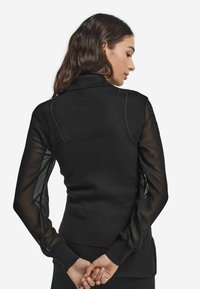 Czarny top z dopasowanym dzianinowym korpusem i przejrzystymi czarnymi rękawami. Posiada kołnierzyk i delikatne przeszycia z tyłu.