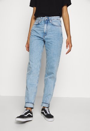 Frau trägt hellblaue High-Waist-Jeans, an den Bündchen aufgerollt, und schwarze Sneakers mit weißen Schnürsenkeln, steht vor einem weißen Hintergrund.