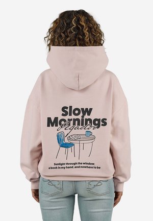 Person iført en lyserød hættetrøje med teksten "Slow Mornings", blå stol, bord med illustration af bog og kop på bagsiden.