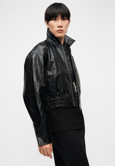 Νεαρός με κοντά μαύρα μαλλιά που φοράει μια υφασμάτινη μαύρη δερμάτινη ζακέτα με ψηλό γιακά και φερμουάρ πάνω από μαύρα ρούχα.