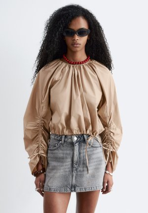 Femme aux longs cheveux bouclés portant des lunettes de soleil noires, un chemisier beige froncé avec des cordons, un collier de perles rouges, une jupe en jean grise et des bracelets.