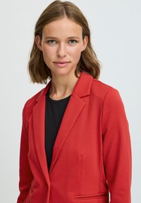 Röd blazer med skräddarsydd passform, enknapprad, notch-slags, och två framfickor, bärs över en svart topp. Slät tygstruktur.