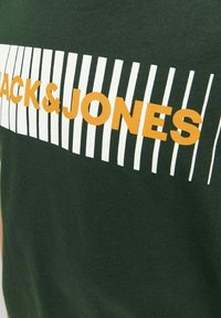 Grøn bomulds T-shirt med et hvidt og orange stribet grafisk design, hvor teksten "JACK & JONES" er iøjnefaldende præsenteret. Blød tekstur.