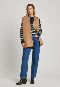 Kamelfärgad ullväst med knappstängning, bärs över en randig tröja i marinblått och kräm. Matchas med högmidjade blå jeans och bruna ankleboots.