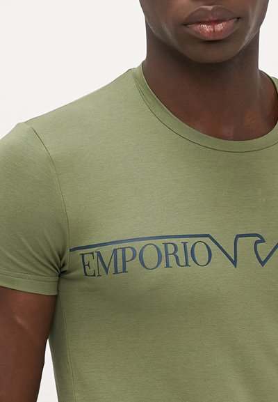 Olīvu zaļa kokvilnas t-krekls ar noapaļotu kakla līniju un īsām piedurknēm, uz krūtīm ir tumši zila "EMPORIO" logo.