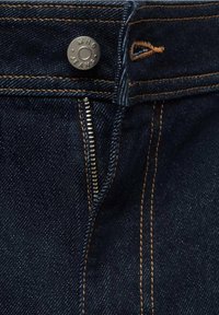 Gros plan sur un jean en denim bleu foncé avec des coutures orange, un bouton métallique gravé "MNG JEANS" et une braguette partiellement ouverte.