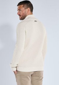 Mann mit kurzem, dunklem Haar und Bart, der einen cremefarbenen, gerippten Pullover und beige Hosen trägt, seitlich steht und die Hände in den Taschen hat.
