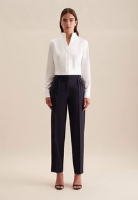 Blusa corta con colletto bianco e dettagli a bottoni, abbinata a pantaloni navy a vita alta con pieghe, tessuto liscio e vestibilità sartoriale.