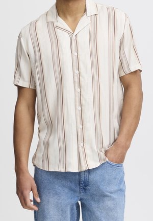 Mężczyzna noszący krótki rękaw, zapinaną na guziki koszulę z pionowymi beżowymi i brązowymi paskami, swobodnie wsunięty w niebieskie dżinsy.