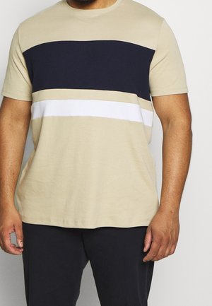Man draagt een beige overhemd met korte mouwen met brede zwarte en witte horizontale strepen over de borst en een zwarte broek.