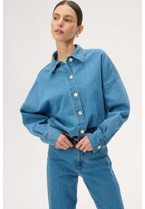 Kobieta poprawiająca czarny pasek, ubrana w luźną niebieską kurtkę dżinsową z białymi guzikami oraz dopasowane niebieskie dżinsy, na tle jasnego, gładkiego tła.