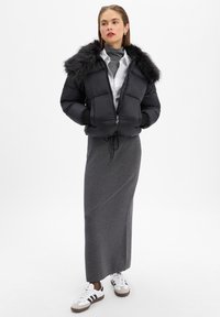 Kobieta ubrana w czarną kurtkę puchową z futrzanym kołnierzem, białą koszulę, szary golf, długą szarą spódnicę i białe trampki w czarne paski.